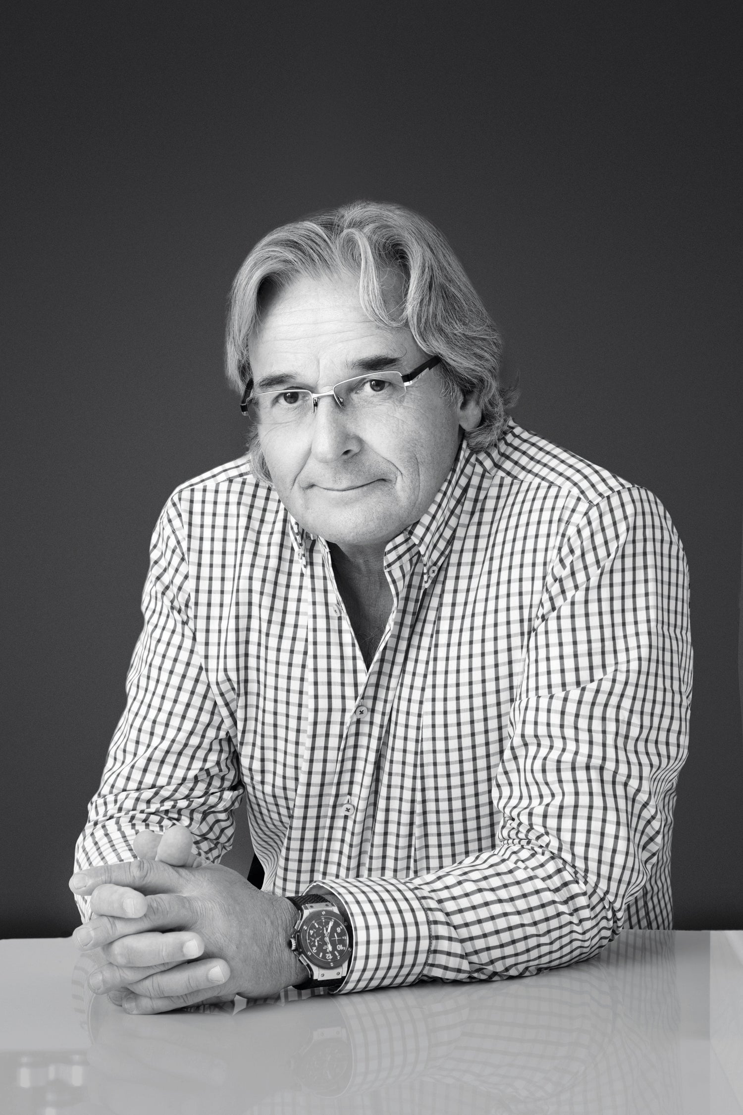 Black-and-white portrait of an older man with glasses and gray hair, wearing a checkered shirt and wristwatch, sitting at a table with his hands folded.