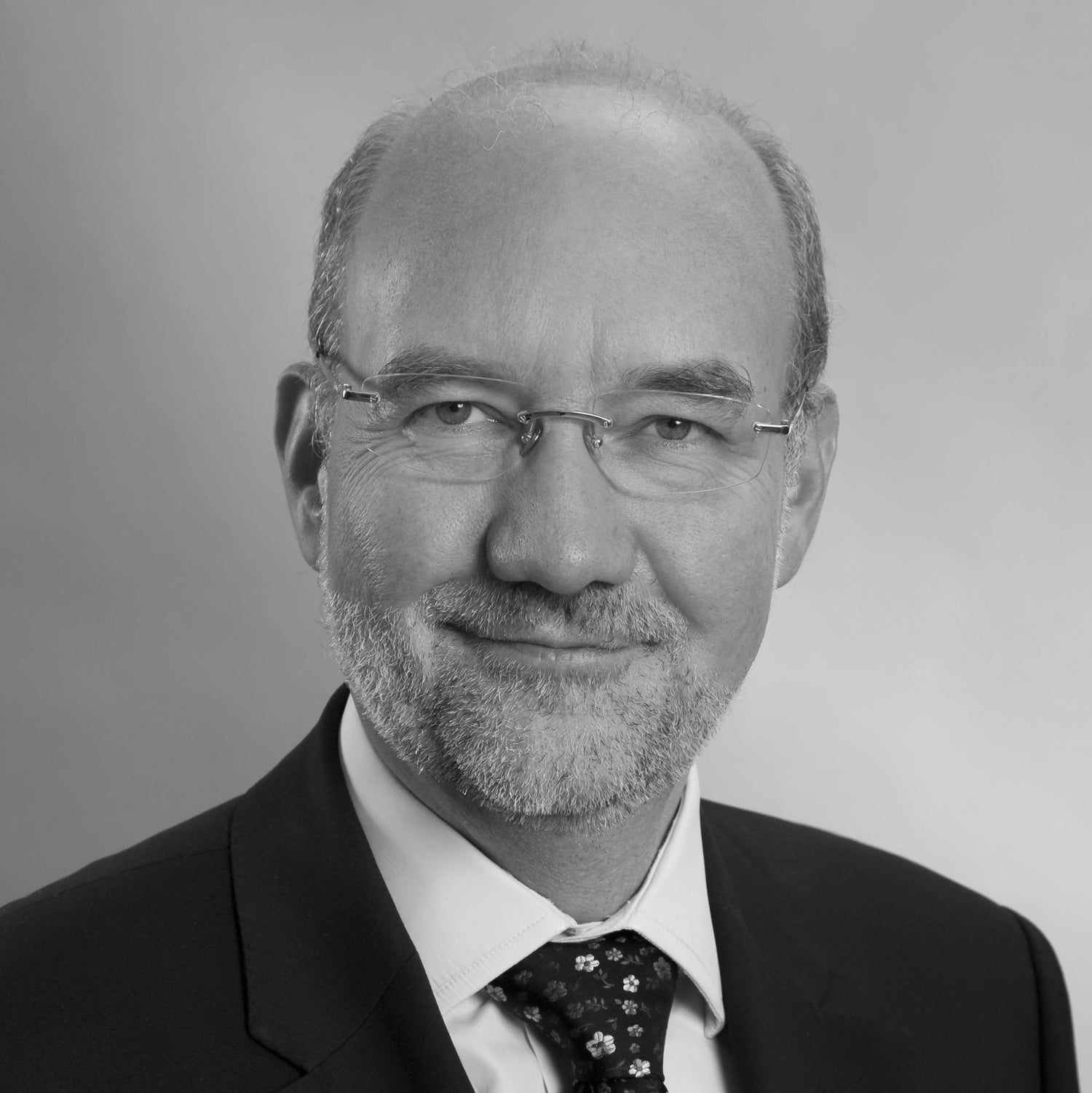 Black-and-white professional portrait of a man with glasses, short beard, and receding hairline, wearing a suit, white shirt, and patterned tie.